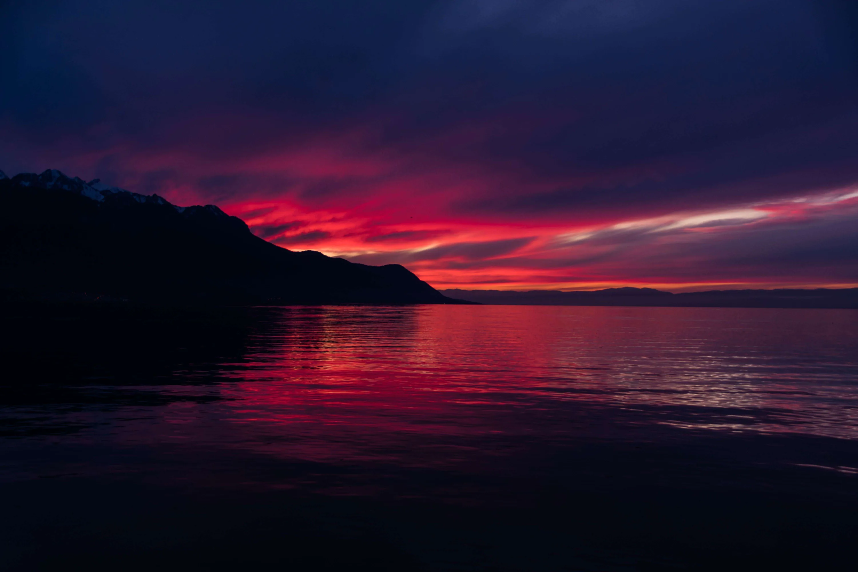 A black mountain slope falls from the left into a lake while deep red cloud burns along the horizon, its colour smearing down the rippled water.