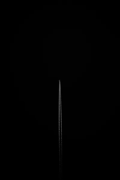 A single thin ridged spike stands upright in the lower half of a tall black frame, catching a narrow highlight down one side with a dim round glow behind it.