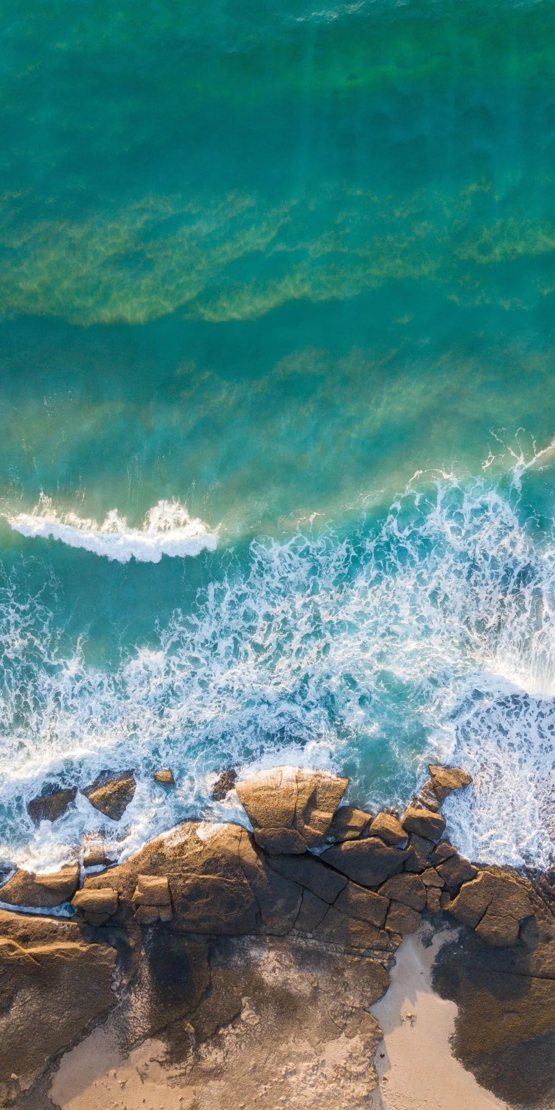 Straight-down view of surf foaming over brown rocks, the green water sliding to deeper teal along the top of the frame.