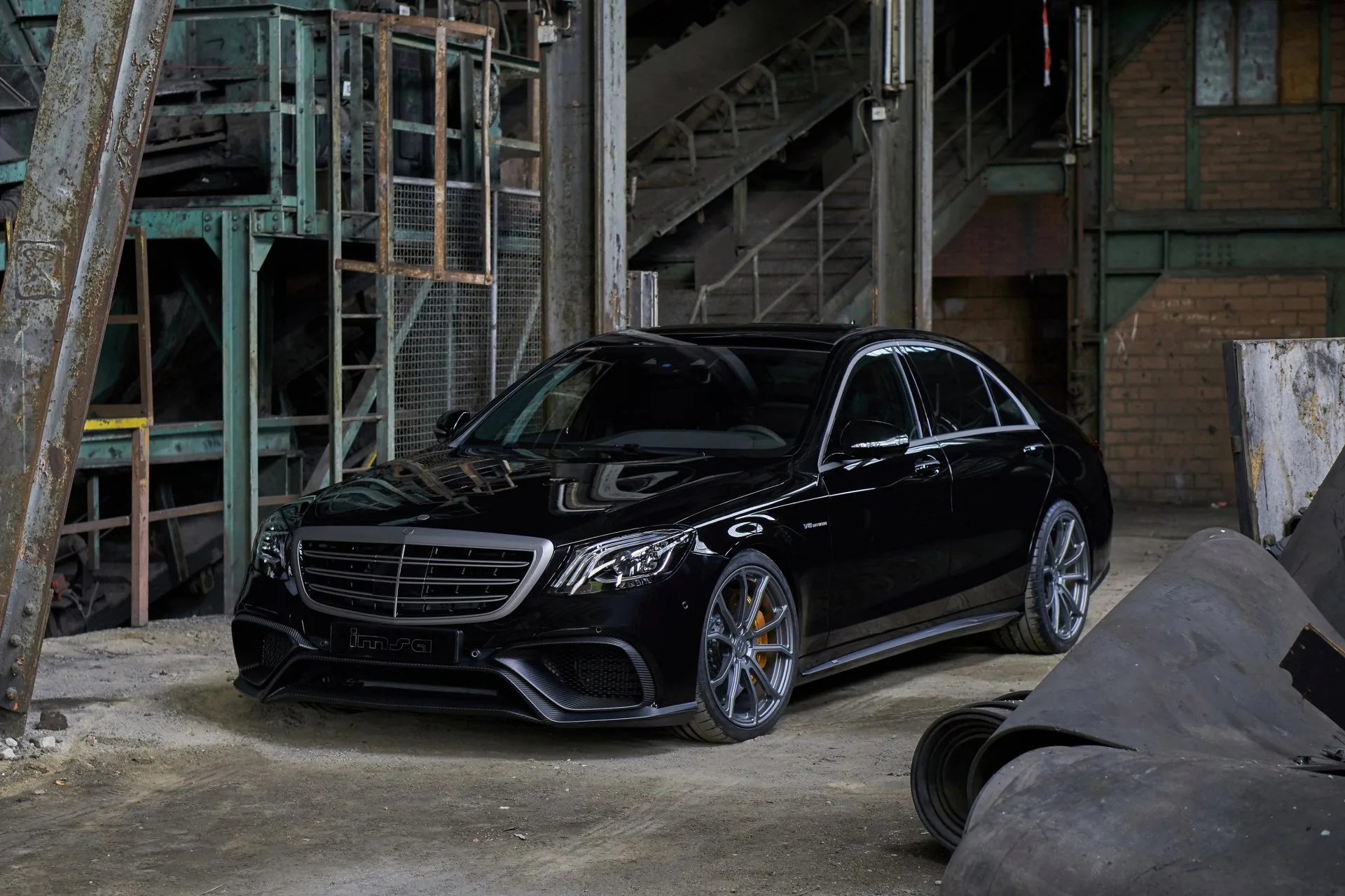 An AMG-styled S-Class parked on the concrete floor of a derelict factory, grey daylight washing in from the left past rusted stairs.