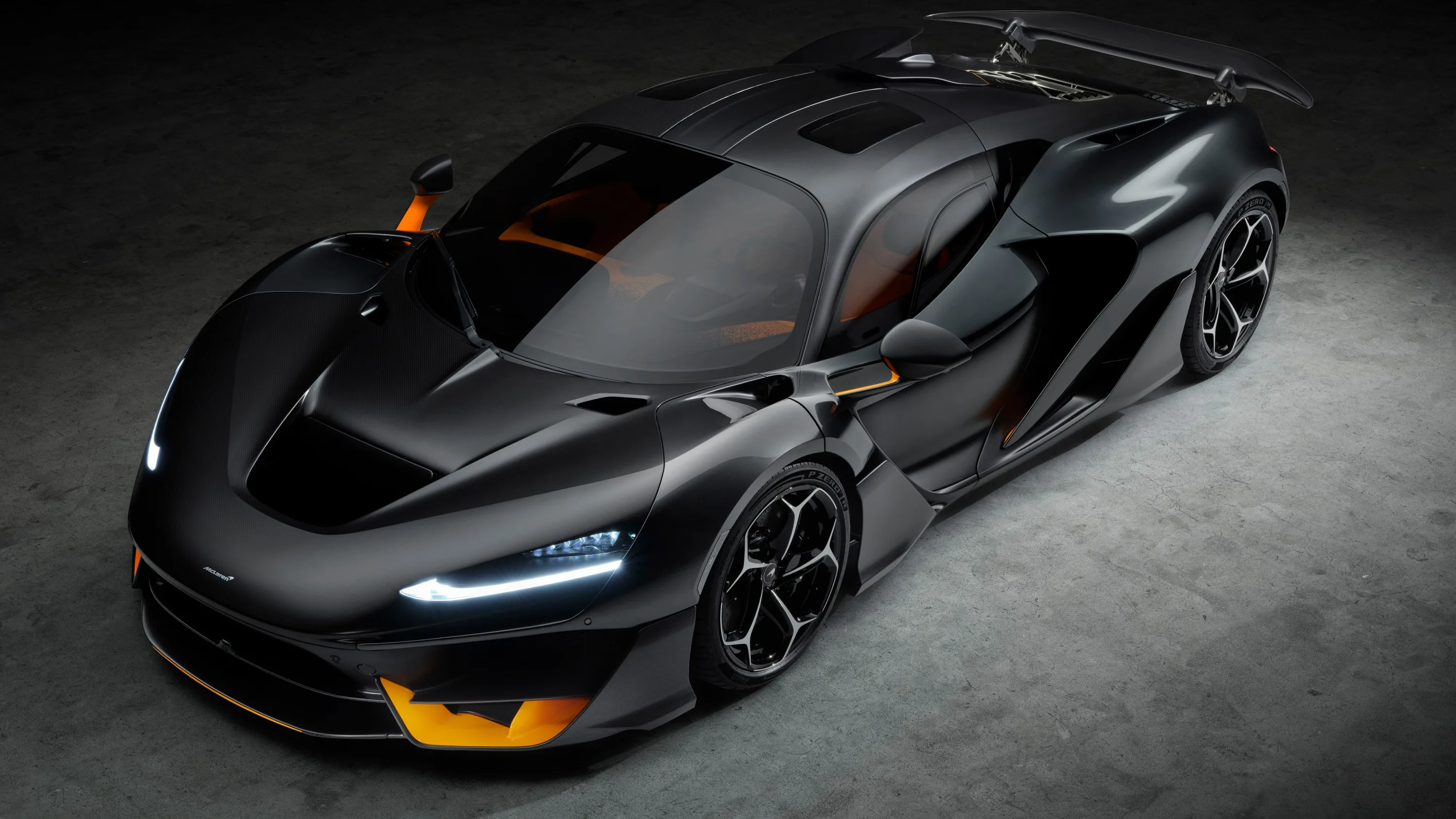 A bare carbon fibre hypercar photographed from above in a grey studio, orange splitter and vents breaking up the dark weave.