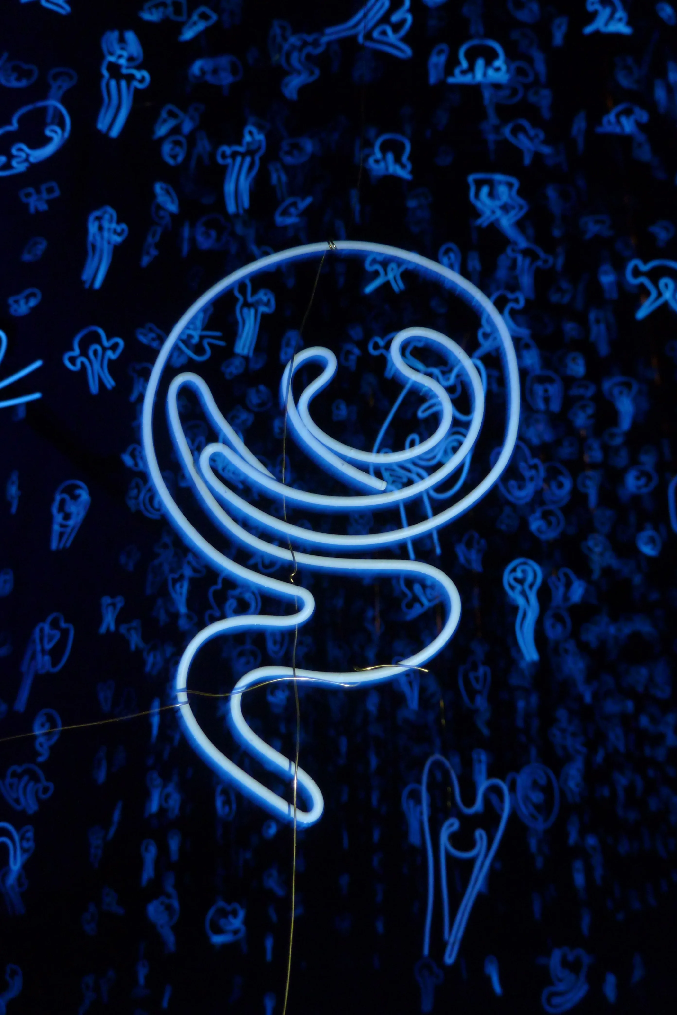 A looping tube of blue light coils across a wall of faint sketched figures, its glow strongest in the centre of the frame.