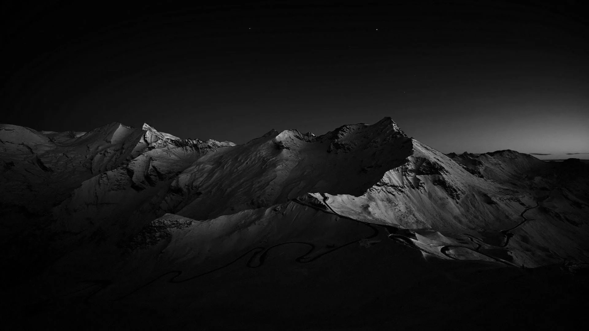 A snow-covered ridge lit from the left in near-darkness, a thin band of dusk light still showing above the far peaks.