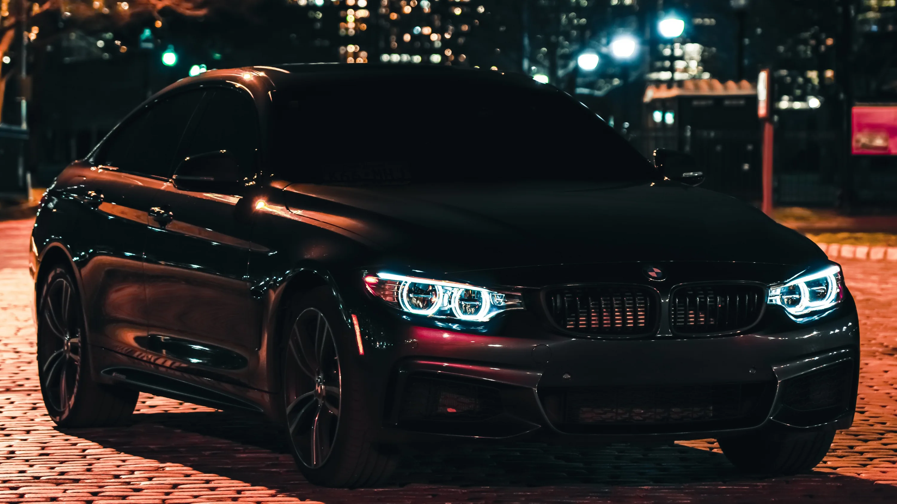 Front three-quarter on cobbles, corona headlights lit and a long string of out-of-focus office windows and streetlights stretching away behind the roofline.
