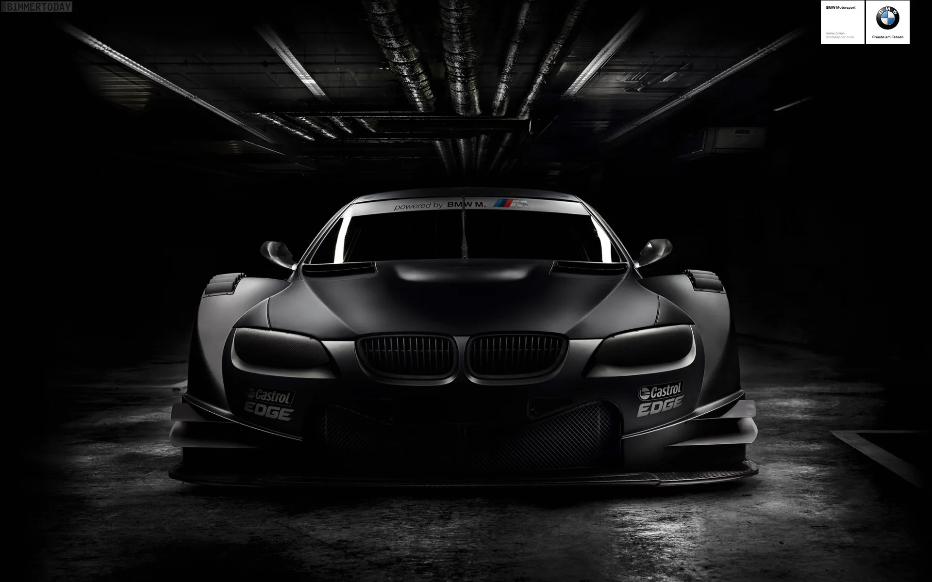 Wide-arch BMW race car facing the camera in a tunnel, strip lights streaking away overhead and monochrome grading pushing everything but the kidney grilles into grey.