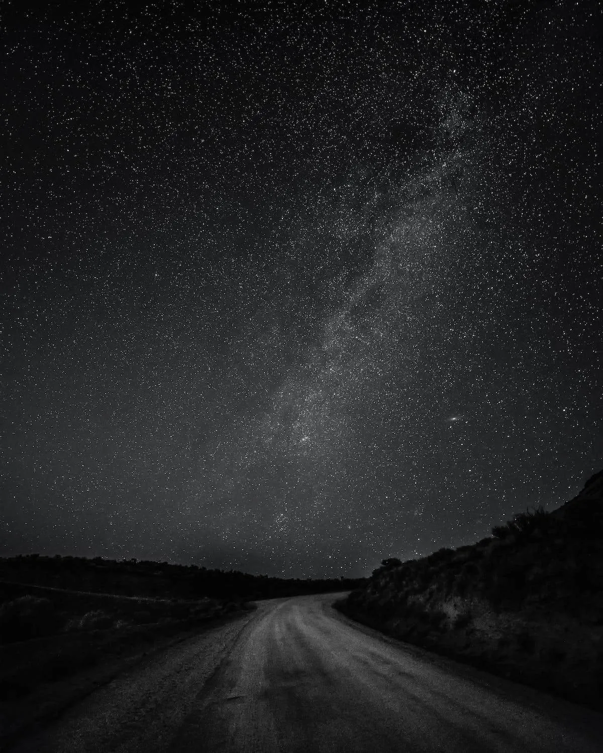 A gravel road runs out of the bottom corner into black hills, the Milky Way climbing diagonally overhead in grainy monochrome.