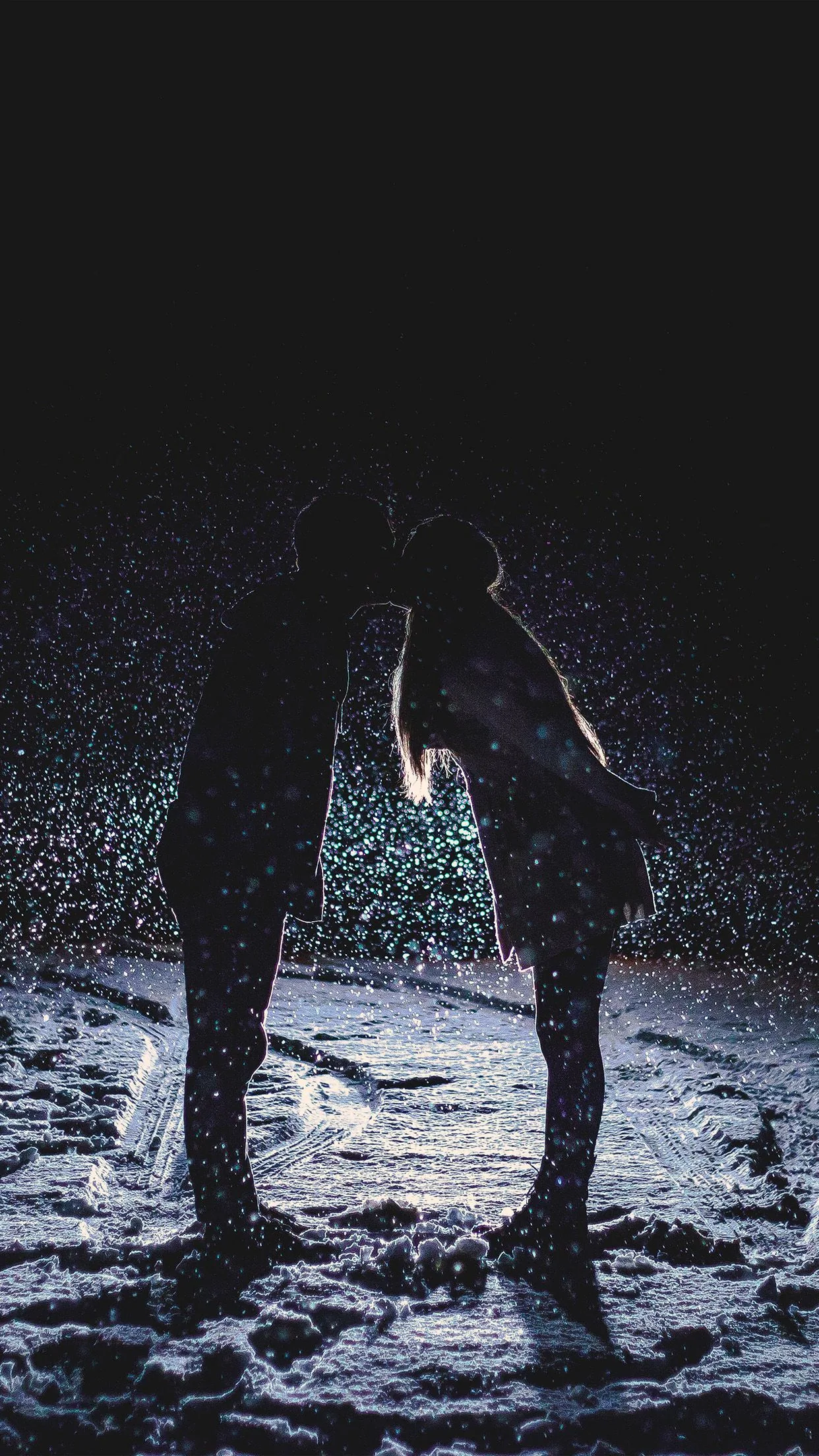 Vertical night shot of two silhouettes leaning together over a lamplit path, snow specks drifting everywhere; the top third stays dark enough for a phone clock.