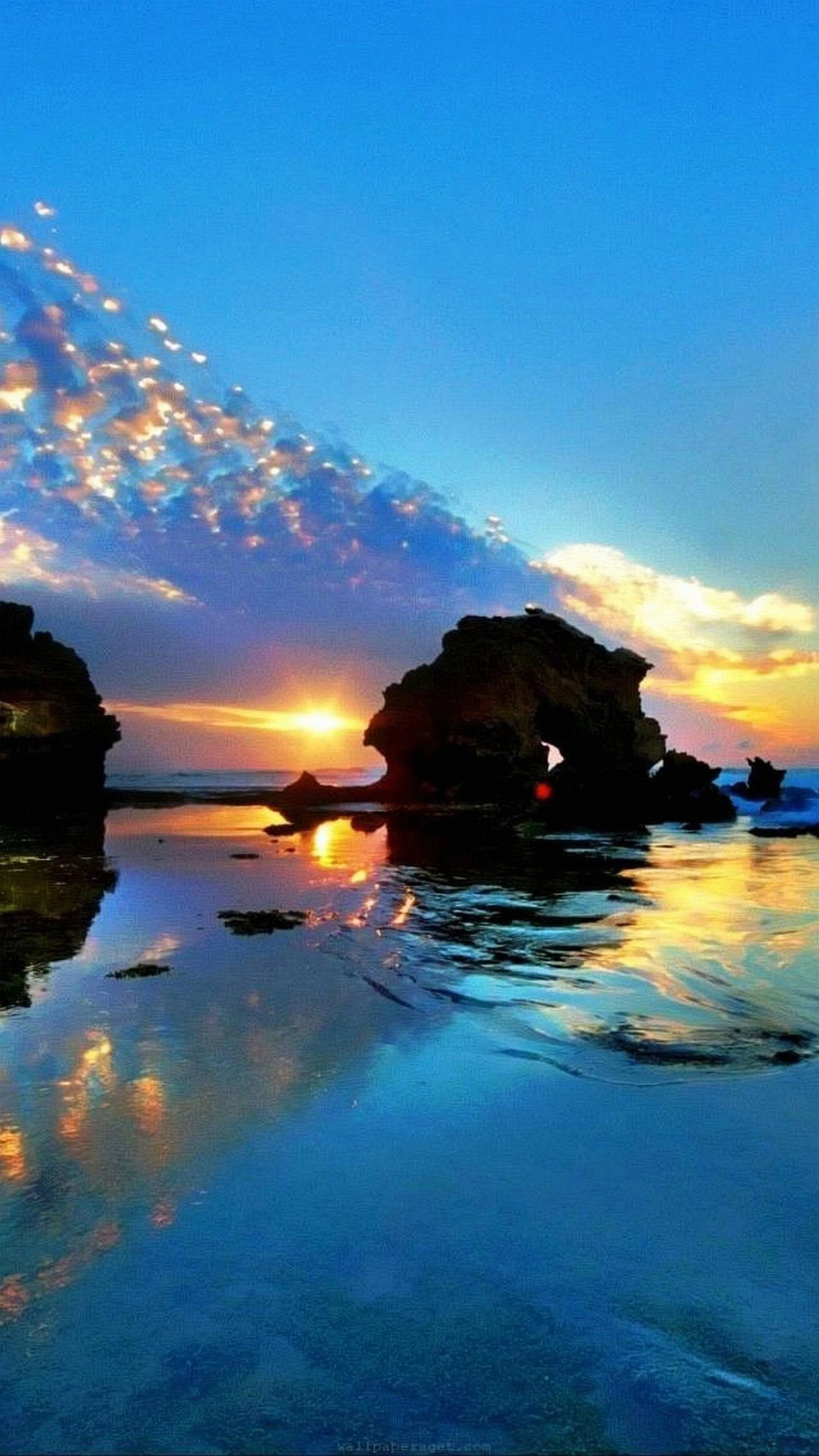 The sun drops between two sea stacks while a trail of golden sparkles climbs the upper-left sky, wet sand mirroring the blue and amber below.