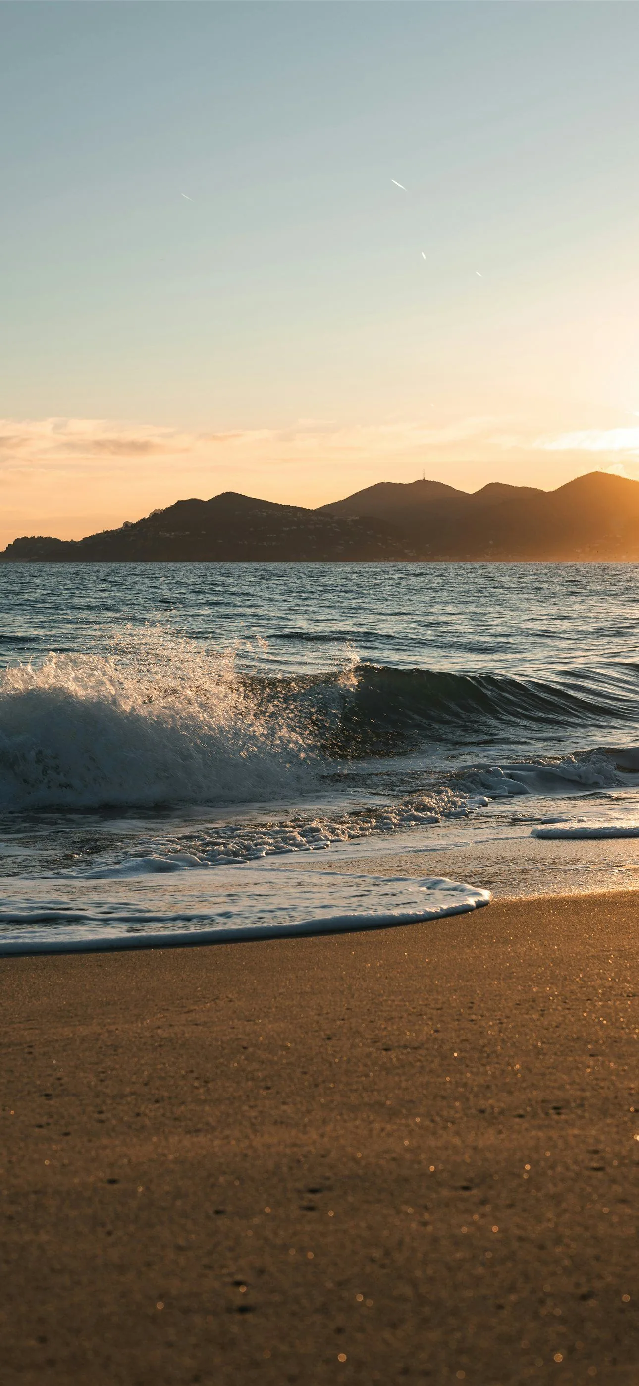 A small wave throws up spray against distant mountains, warm low light grazing the sand that fills the lower third of the frame.