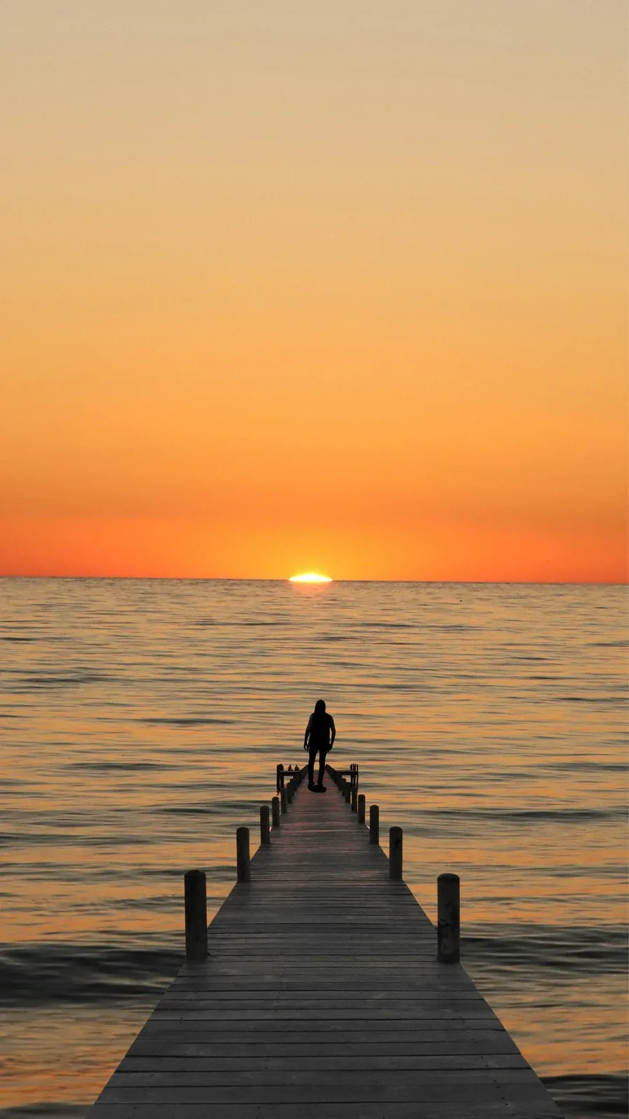 A figure sits at the end of a long wooden jetty facing the last sliver of sun, the plain orange sky above left empty for a lock-screen clock.