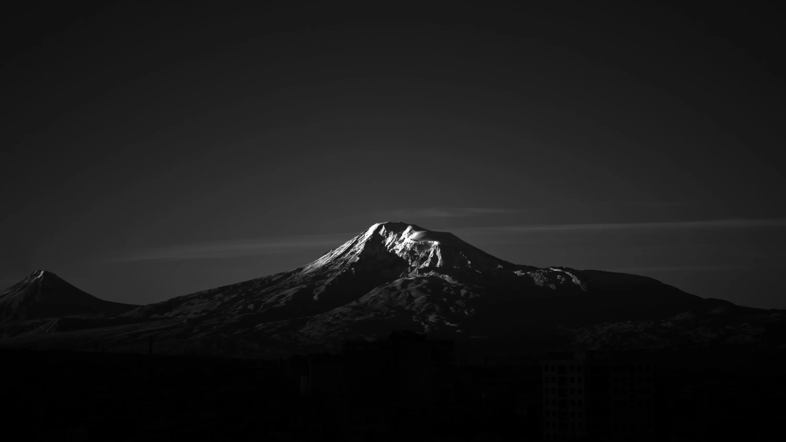 A twin-peaked volcano catches the last light on its snow while the ridges below sink into black, a thin band of cloud crossing the summit.