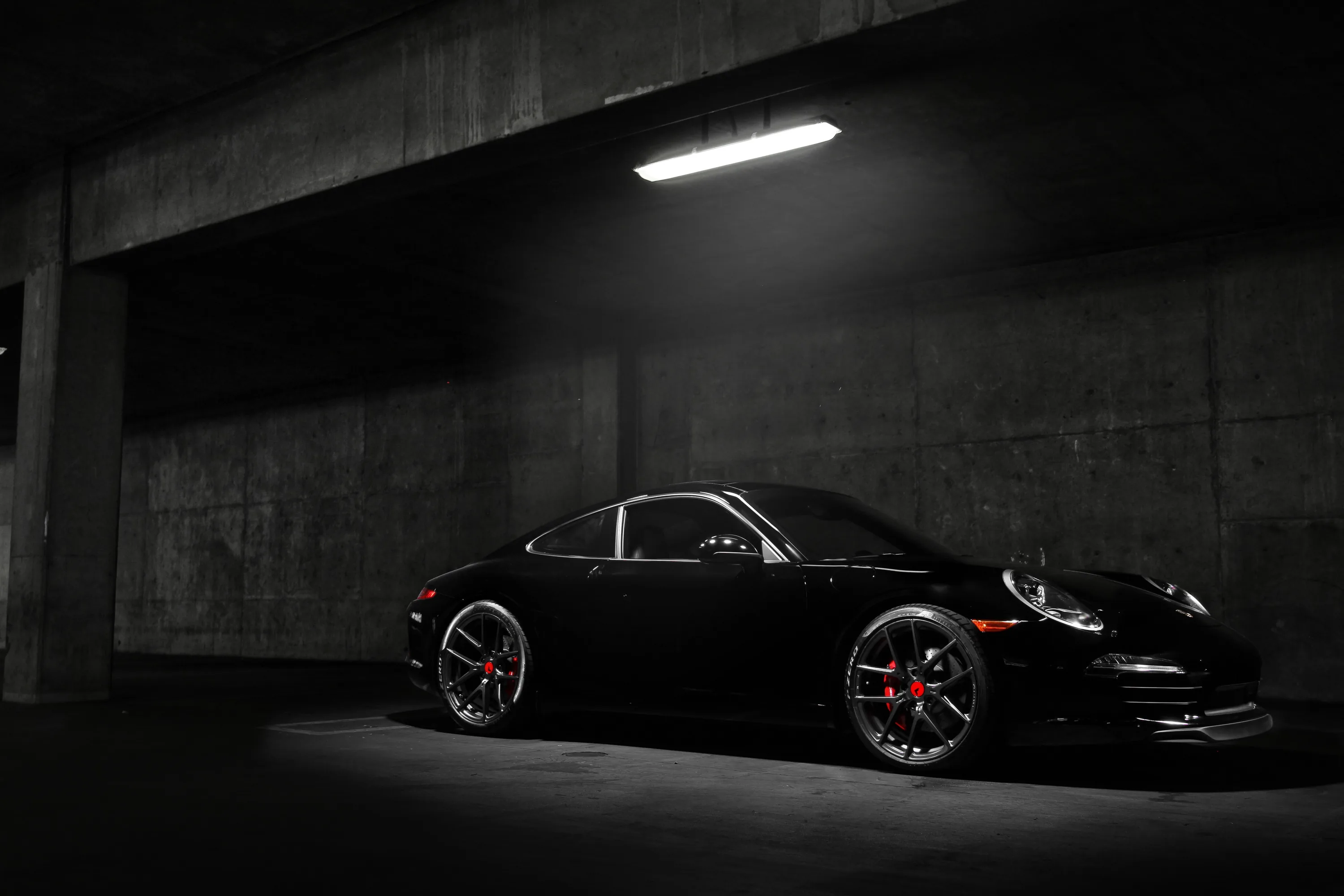 A low 911 waits under a single strip light in a bare concrete garage, red brake calipers showing through the spokes while the left half stays dark.