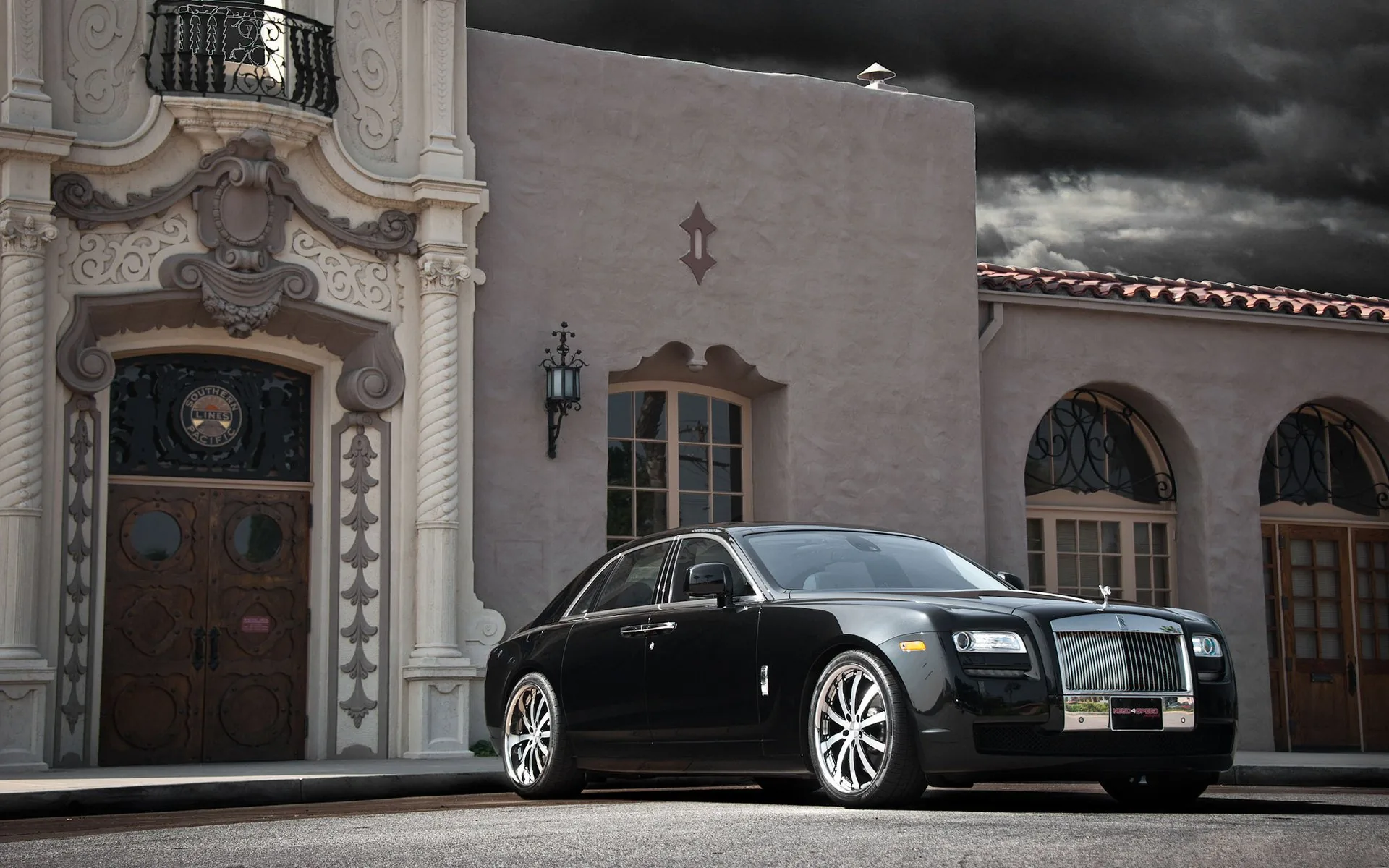 The black sedan sits before a carved Spanish-style entrance, storm clouds stacking over the roofline while the upper sky stays dark enough for a clock.