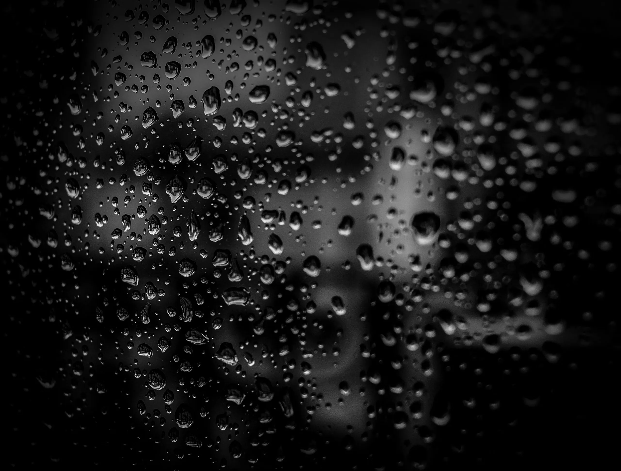 Water beads cling to glass in monochrome, the sharp drops up front giving way to blurred grey shapes somewhere beyond the pane.