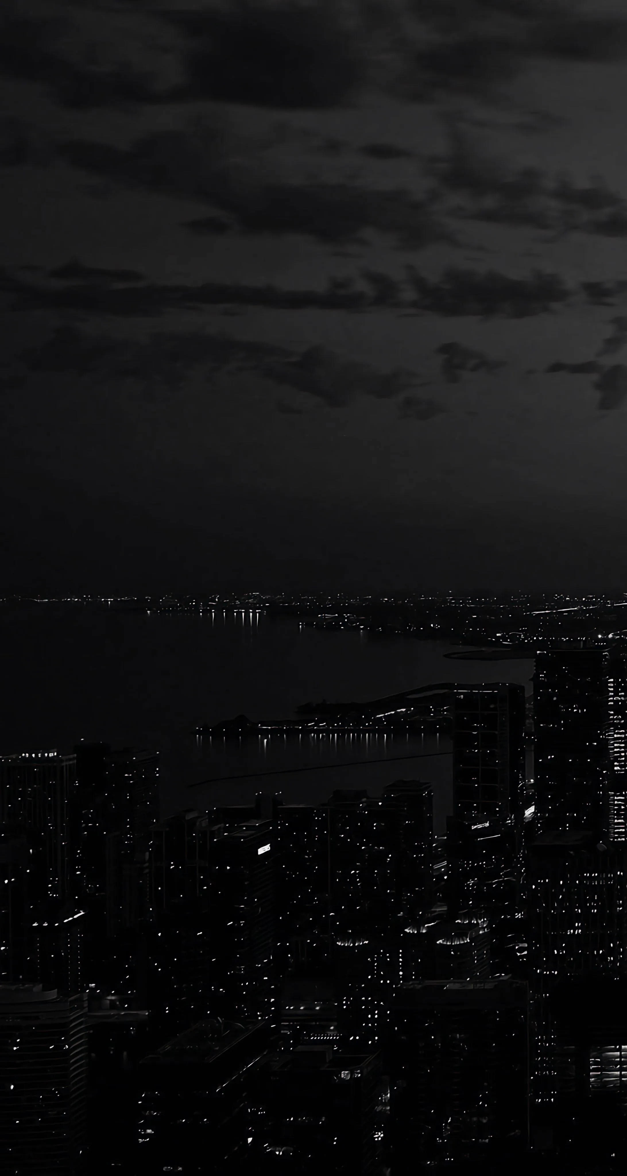 Scattered window lights trace a night skyline along a harbor, with the cloudy sky above left dark enough for the clock to sit clearly.