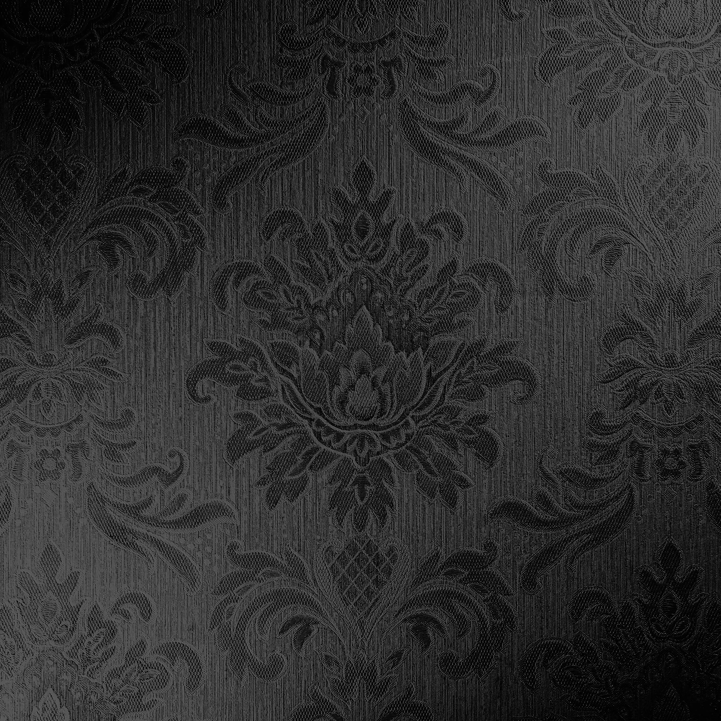 Raised damask motifs press out of a finely ribbed black fabric, the weave catching light near the centre while all four corners fall into shadow.