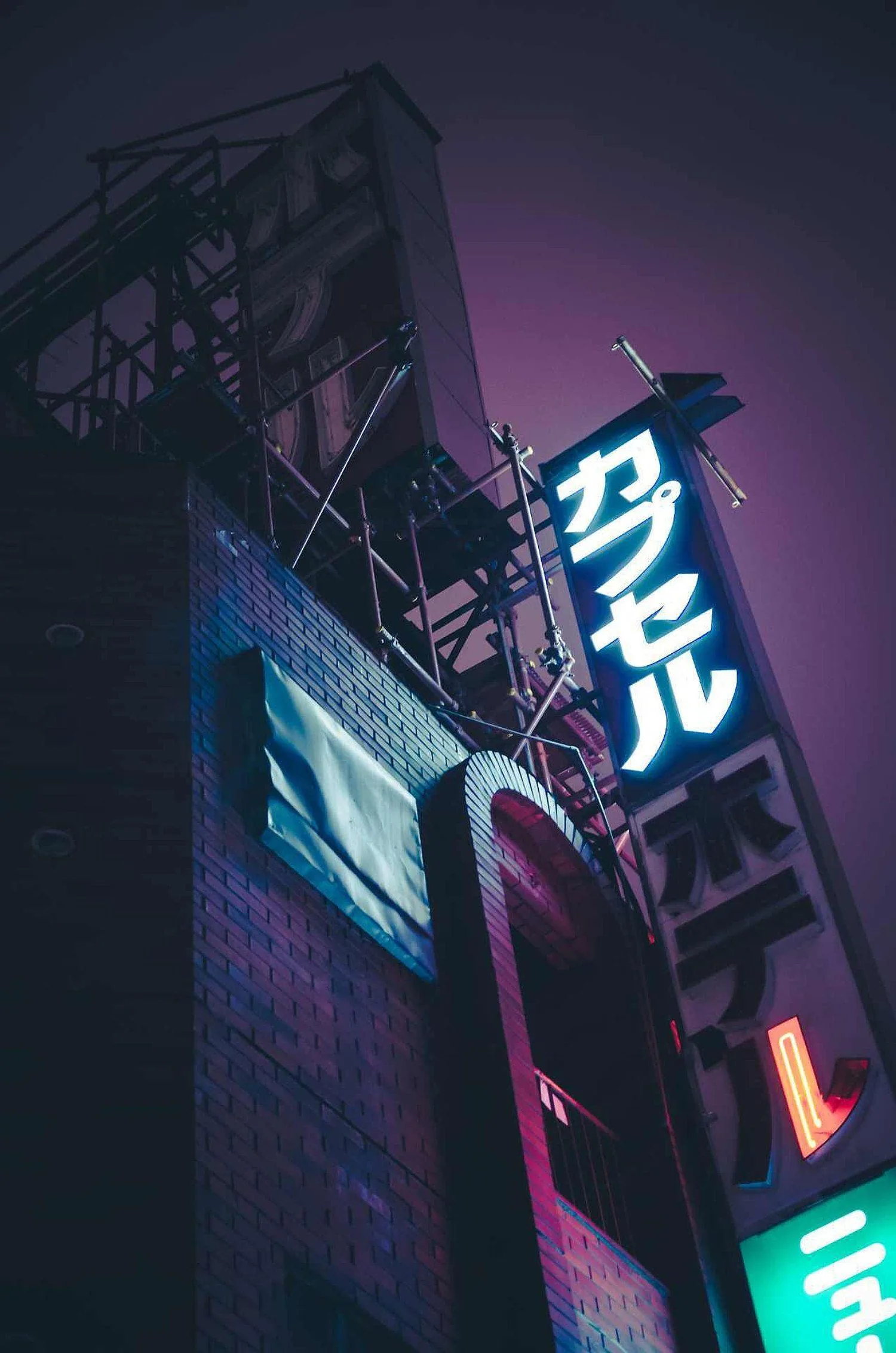 A vertical katakana sign for a capsule hotel glows against purple night haze, the scaffolded building on the left staying almost black.
