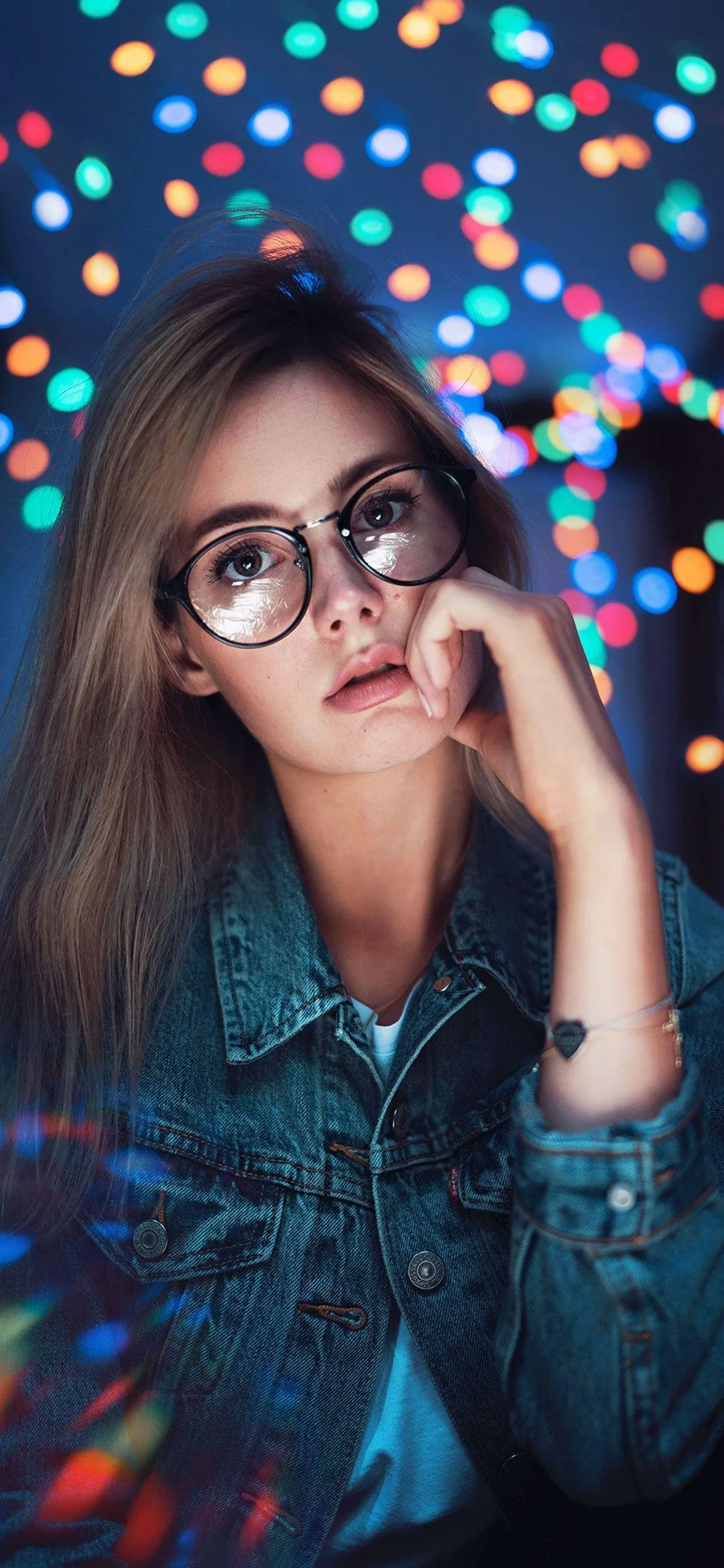 A young woman in a denim jacket rests her chin on her hand, multicoloured fairy lights blurring into bokeh across the upper half of the frame.