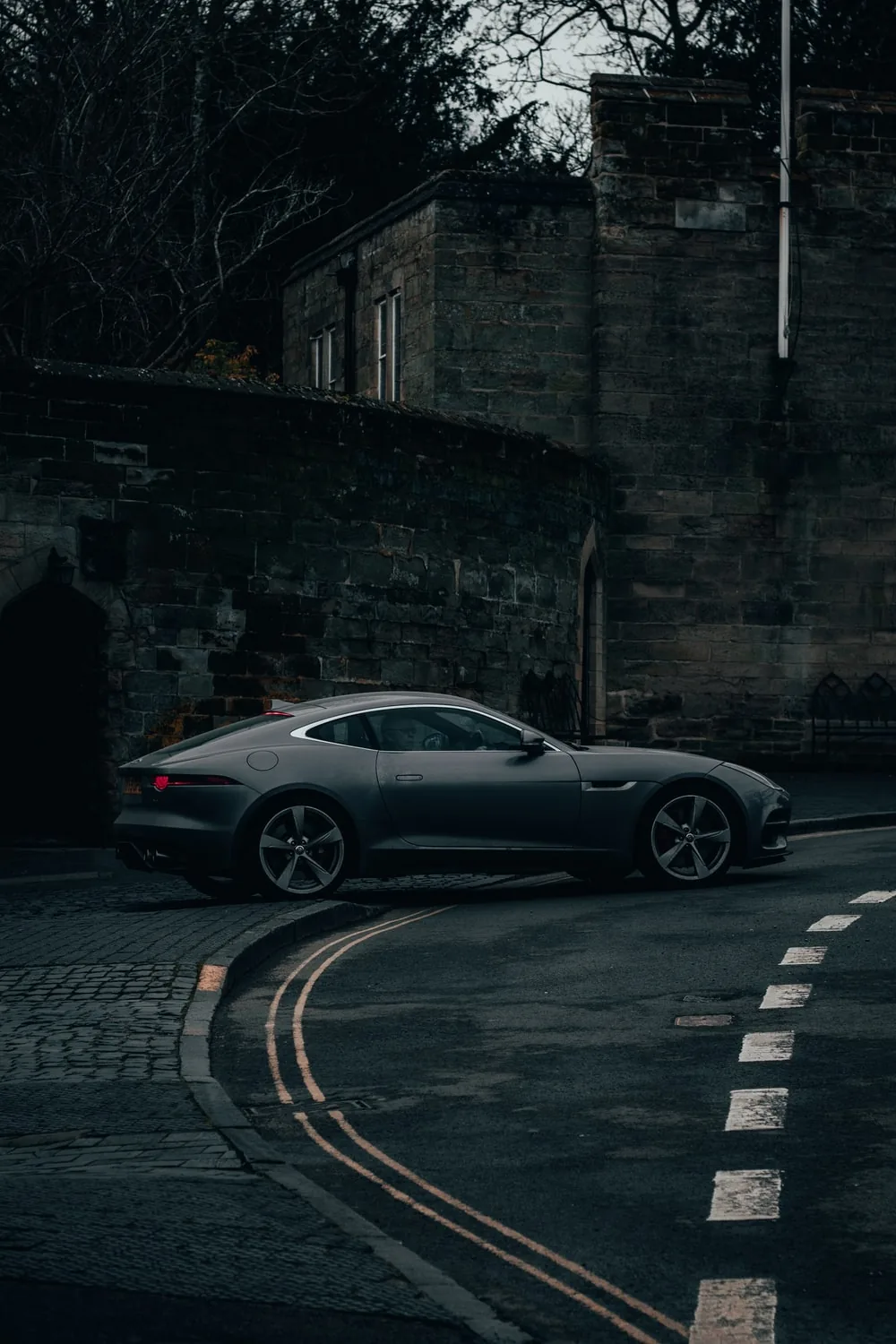 Dusk shot down a narrow lane: a dark coupe follows the double yellow line past an old stone wall, the cobbled kerb and bare trees kept in shadow.