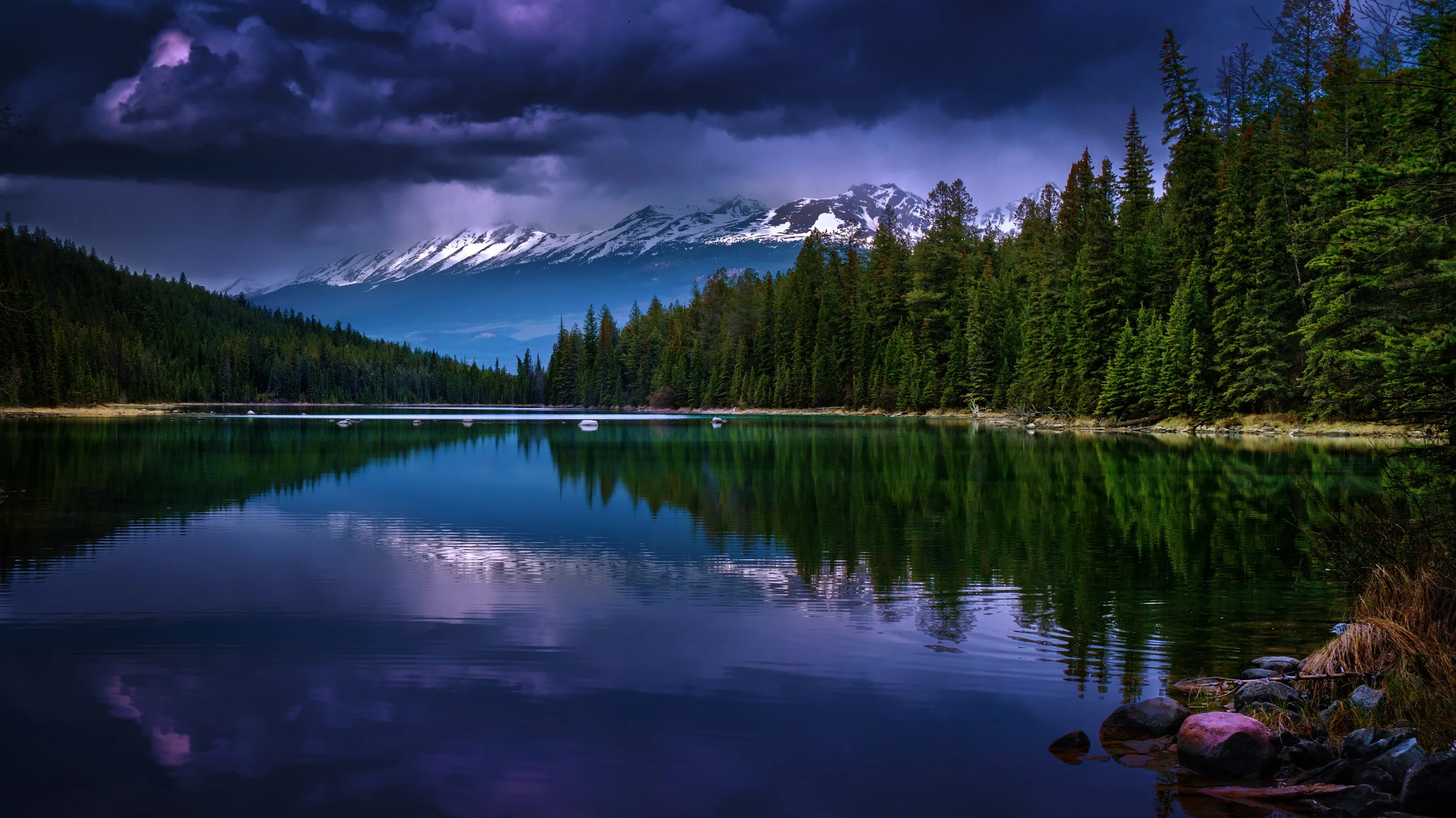Violet storm cloud hangs over snow-dusted peaks while a pine shoreline repeats in the green lake below, boulders breaking the near shore at the lower left.