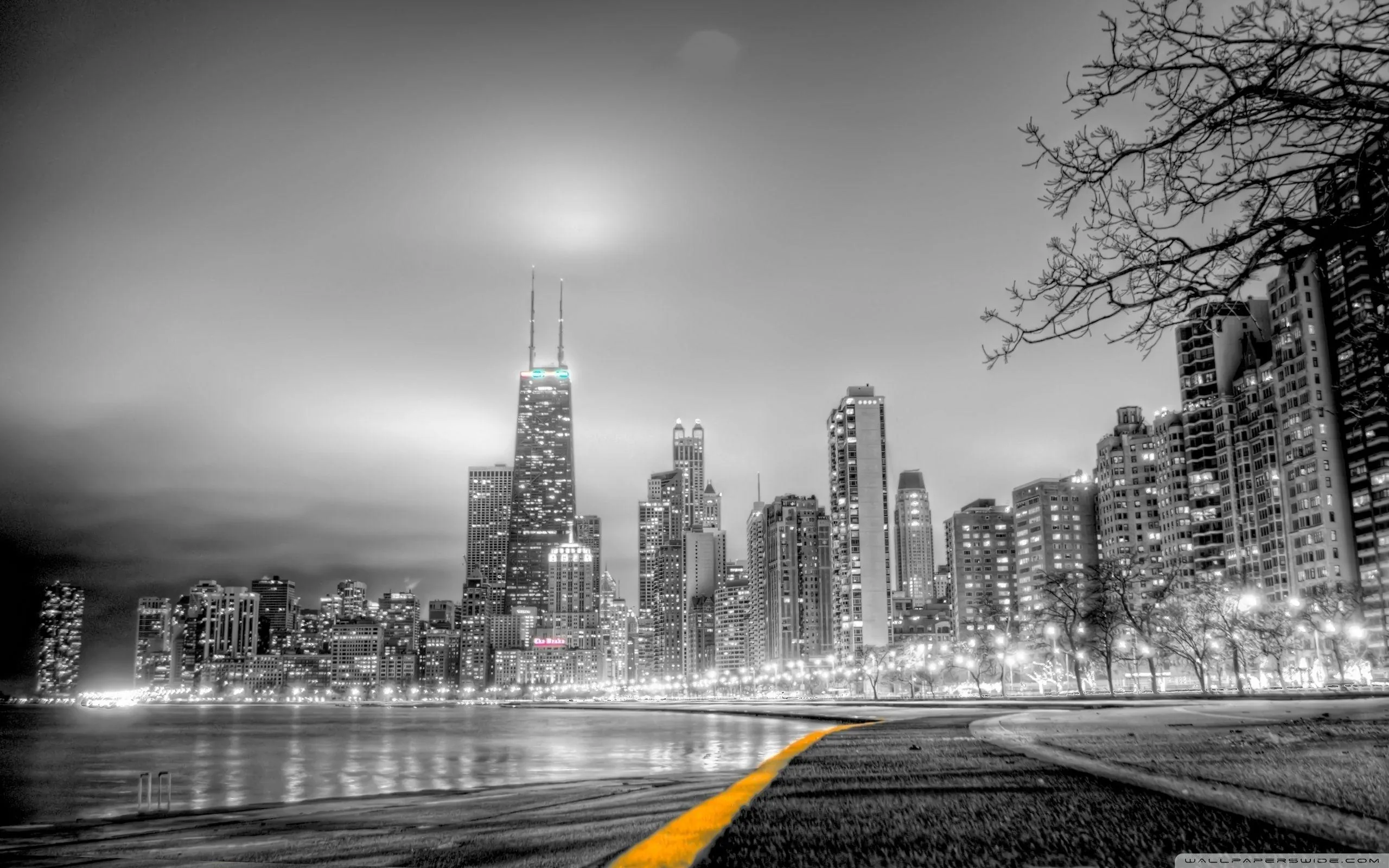 Towers rise in grey haze beyond a curving lakefront road, the painted centre line the one stroke of colour left in the shot.