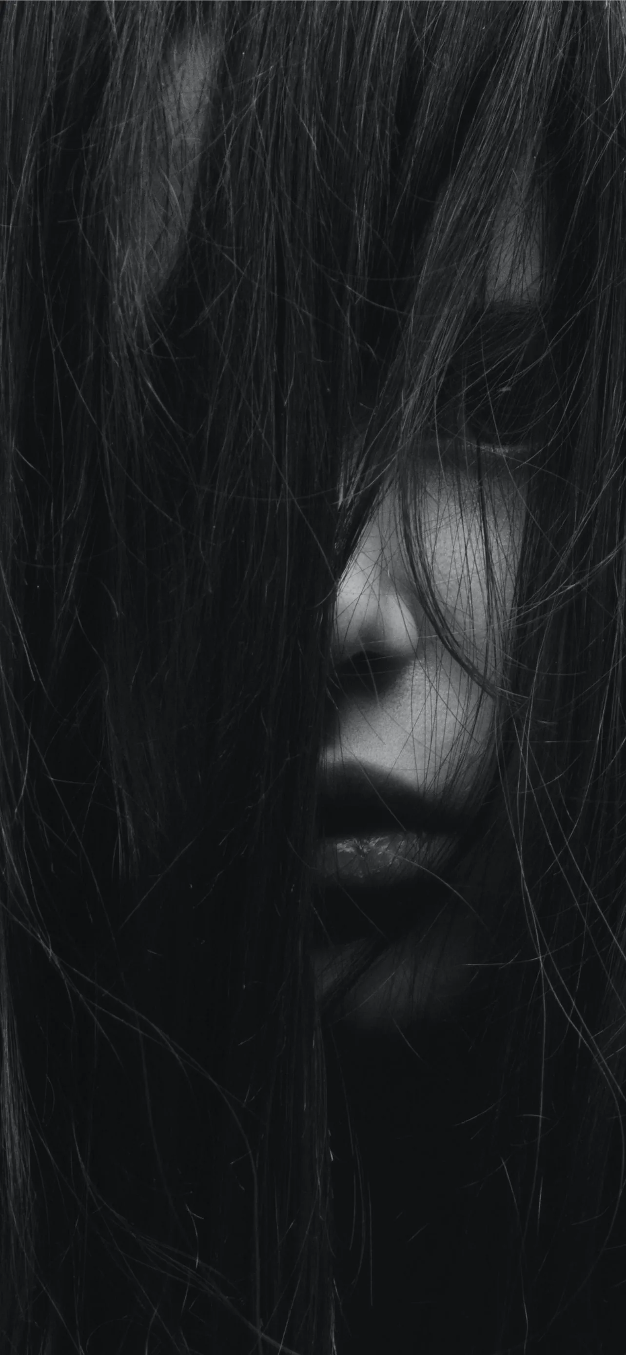 Long strands of hair veil most of the face in grainy black and white, only the lips catching light near the centre of the frame.