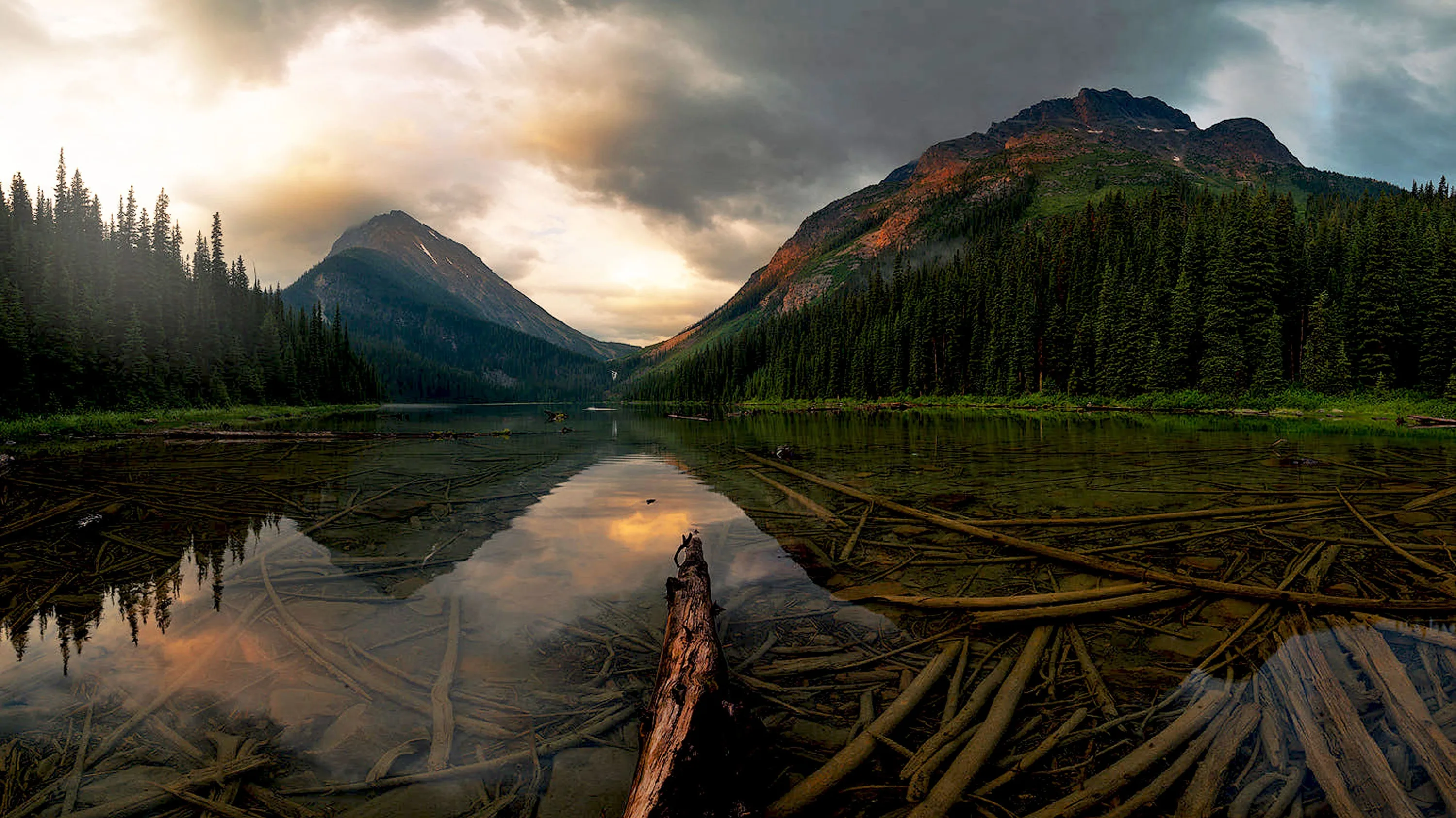 Weathered logs converge from the lower right toward two dark peaks, the last warm light catching the ridge on the right under a breaking storm sky.