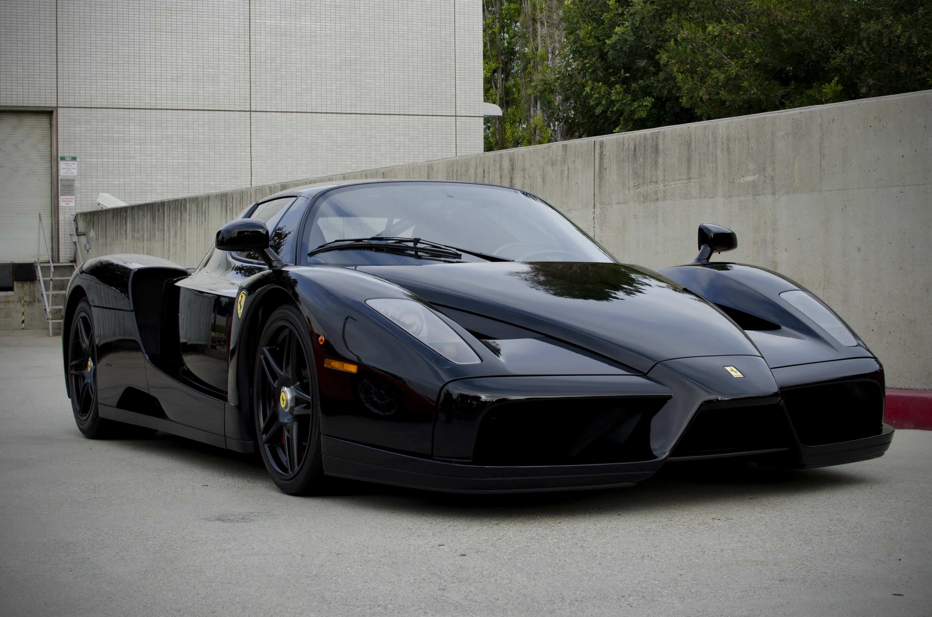 A gloss black Enzo parked on worn asphalt before pale concrete buildings, shot low from its front left, the bright wall keeping the frame's upper edge light.