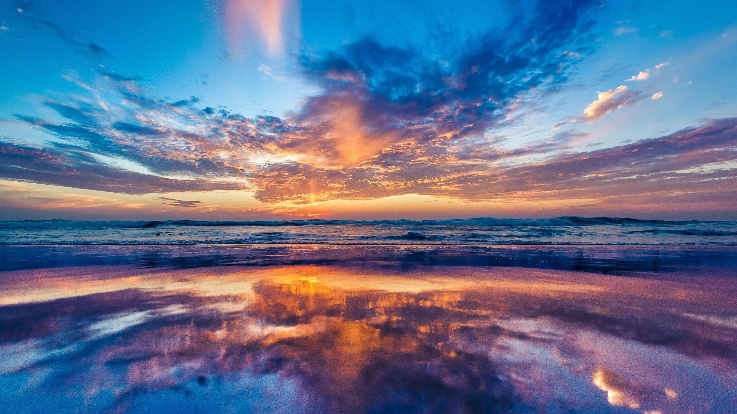 Orange-lit clouds double themselves in the film of water left on the beach, sky and sand meeting in a nearly symmetrical mirror.