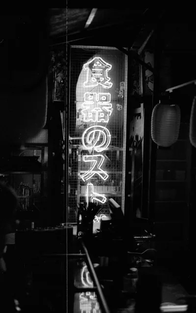 Vertical neon lettering glows down a narrow shop front in grainy black and white, paper lanterns hanging to the right and the foreground counter falling into shadow.