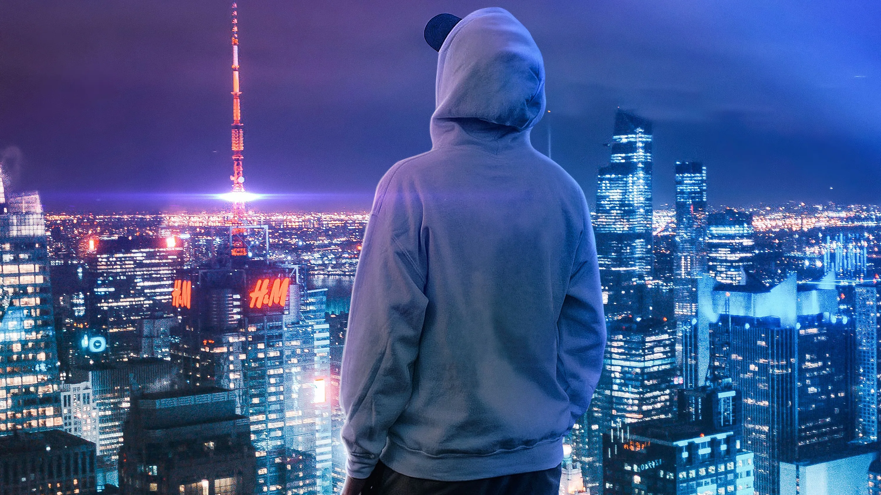 Shot from behind in a pale hoodie, he looks out over a violet skyline with a lit broadcast tower rising on the left.