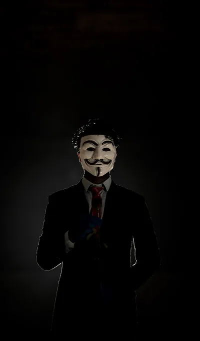 A suited figure in a moustached white mask stands low in the frame, faint warm light from the right, the upper two-thirds left almost black.