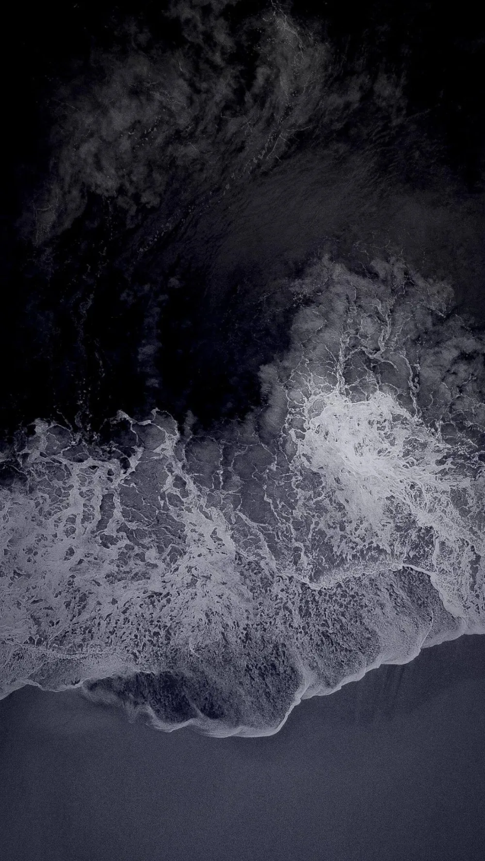 Shot straight down at the shoreline, white surf tears across dark volcanic sand while the deep water at the top stays near black.