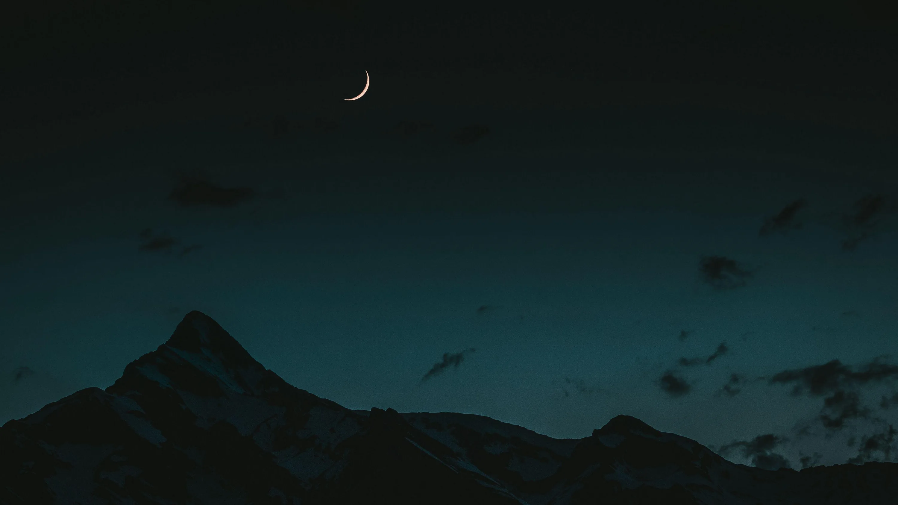 A thin crescent hangs high left of centre in a teal-black sky, the jagged mountain silhouette holding down the lower left corner.