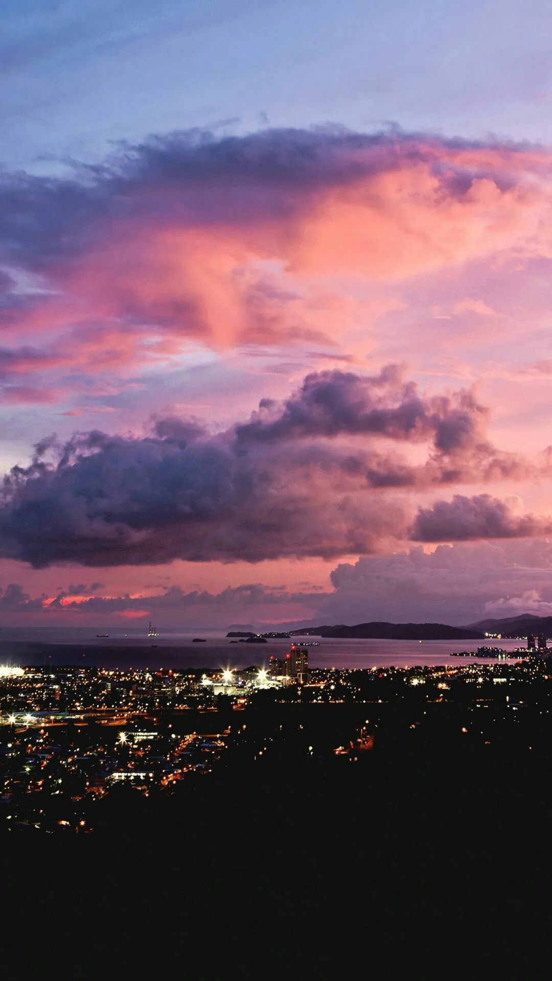 Pink and violet storm clouds stack above a coastal city at dusk, its street lights forming a glowing band across the lower third.