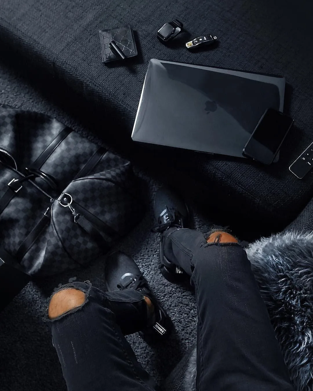 Seen from above, ripped jeans, leather gloves, a checkered duffel and a laptop spread across dark bedding, small keys and a wallet along the top.