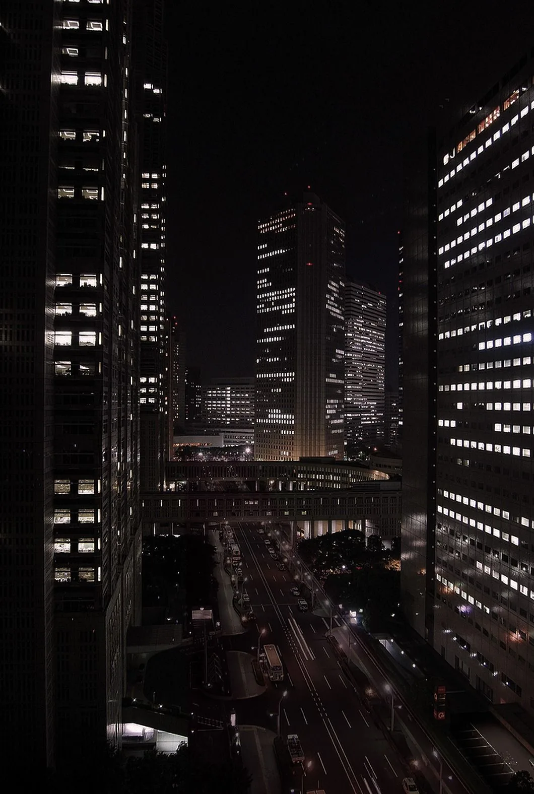 Shot from a high window: glass towers with scattered lit floors on both sides, and far below a multi-lane road smeared with red and white traffic trails.