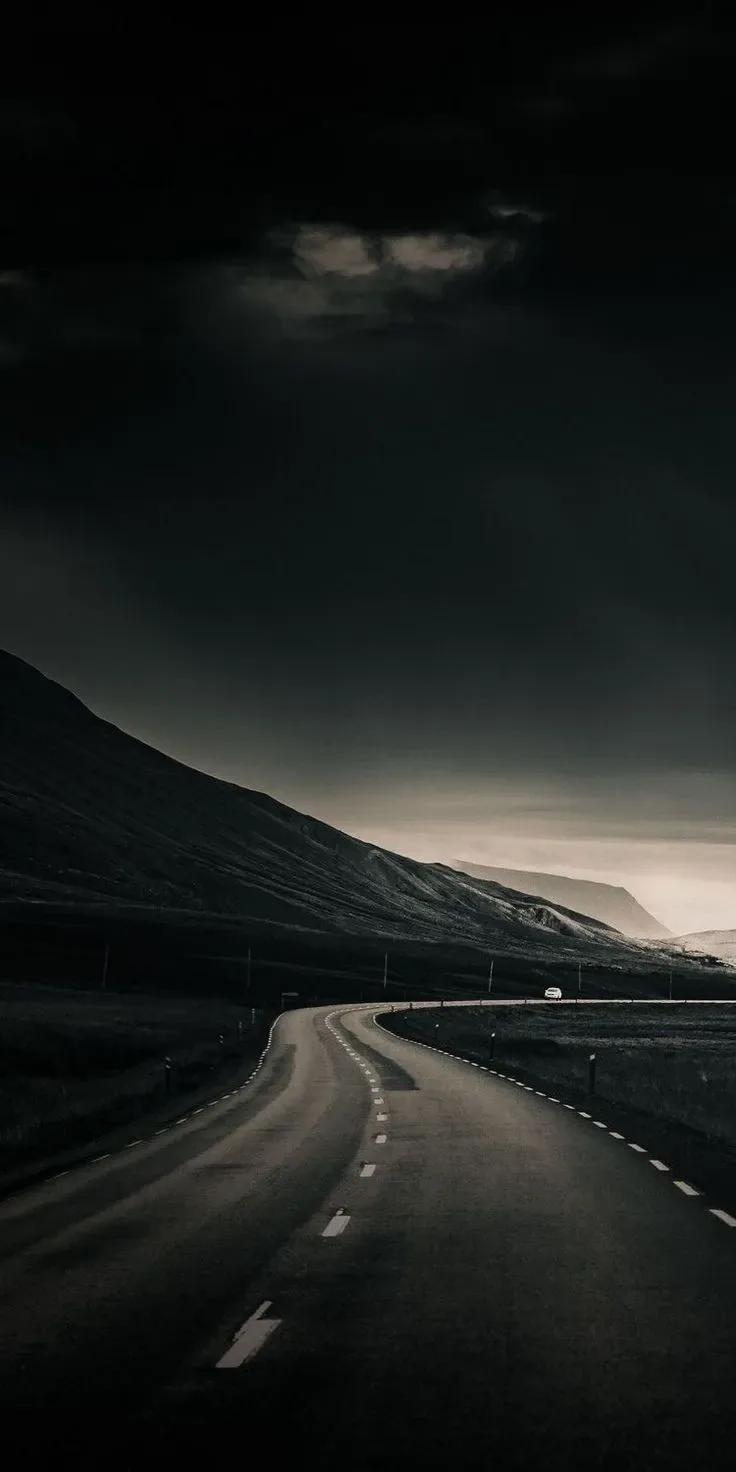 An empty two-lane road bends between dark hills, heavy cloud pressing down and a thin strip of pale light left along the horizon.