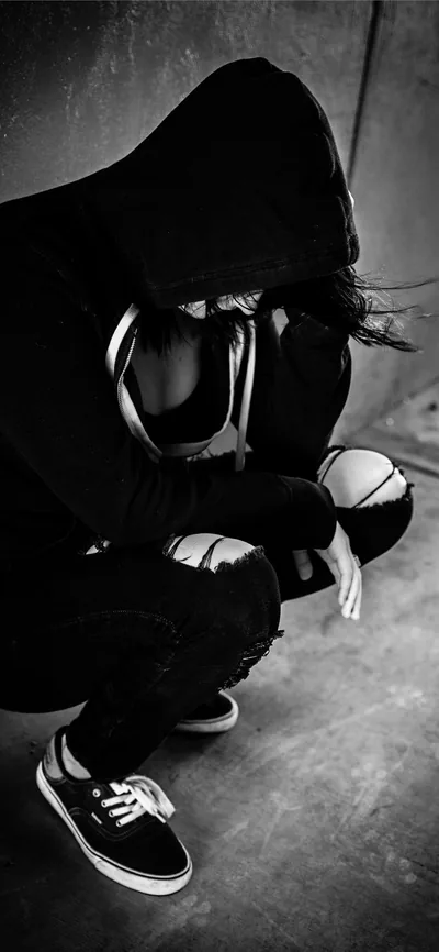 Framed tall for phone screens, a hooded figure crouches low against a grey wall, a checkerboard slip-on at the bottom edge and the face fully lost in shadow.