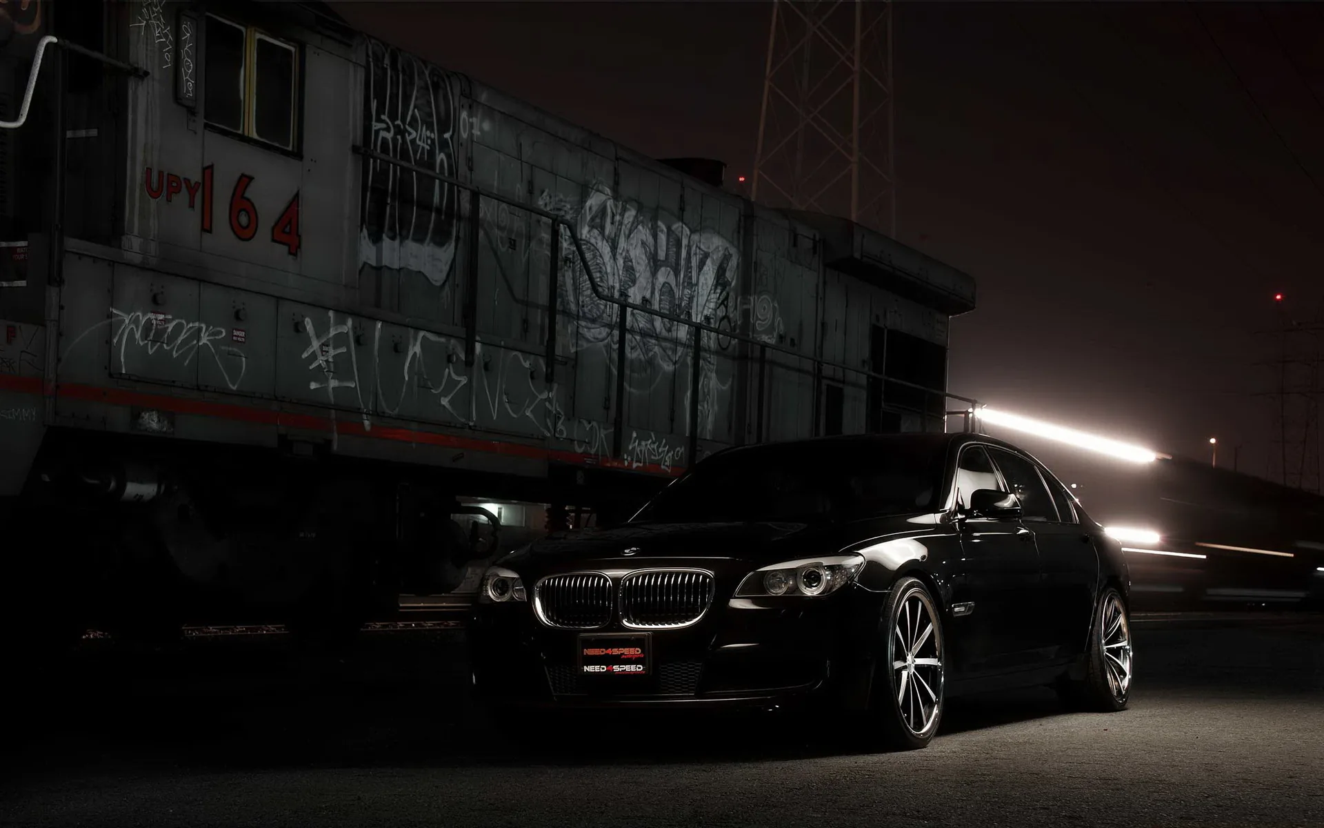 A black 7 Series parked at night next to a graffiti-covered locomotive, a single streak of light passing behind the car's roofline.