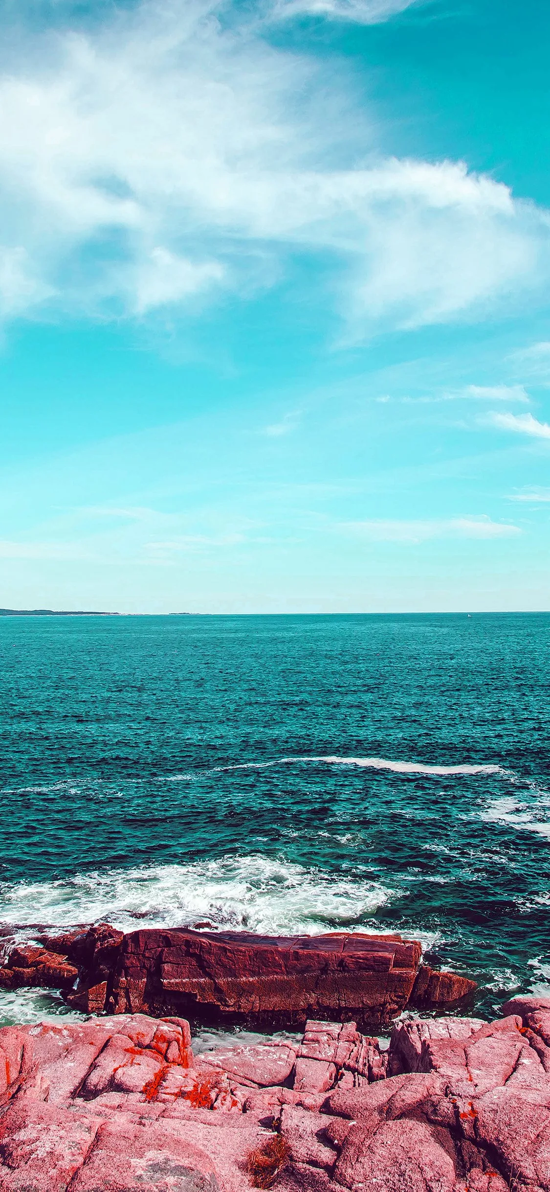 Reddish-pink boulders edge a teal sea under a pale filtered sky, the top half left mostly empty for a phone clock.