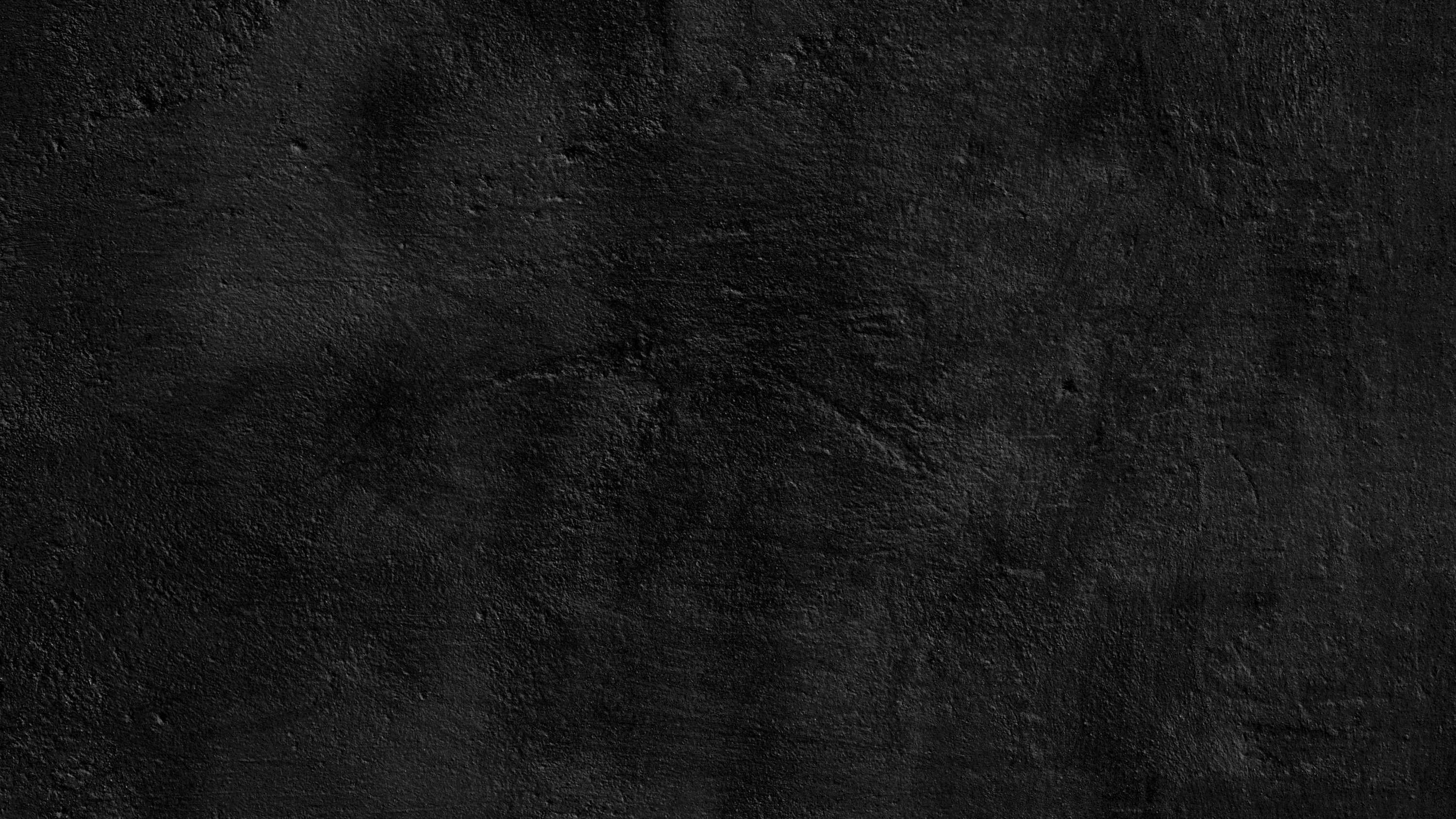 A weathered concrete wall in near black, patchy stains and pit marks spread unevenly, denser along the top edge than the smoother lower half.