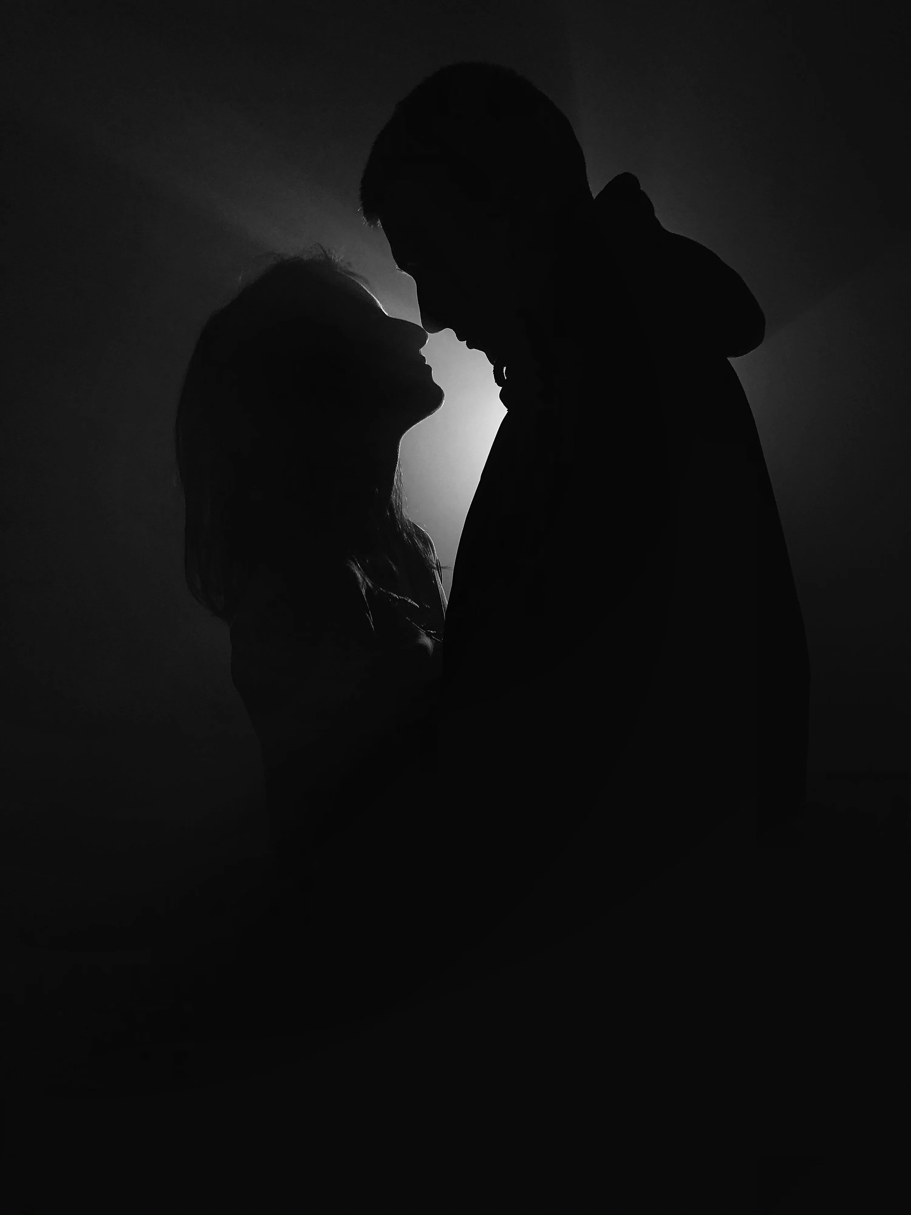 Grayscale silhouettes face each other with only a faint haze of light between their profiles; the hooded figure stands right, and most of the frame stays true black.