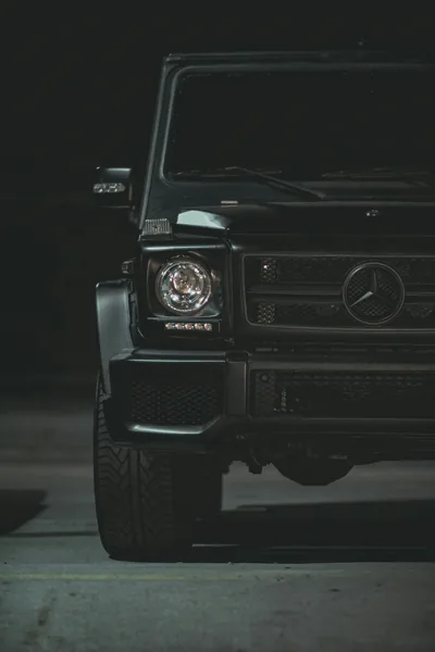 Vertical close crop on the boxy nose of a black G-Wagen, round headlamp and slotted grille lit from the side while the rest sinks into shadow.